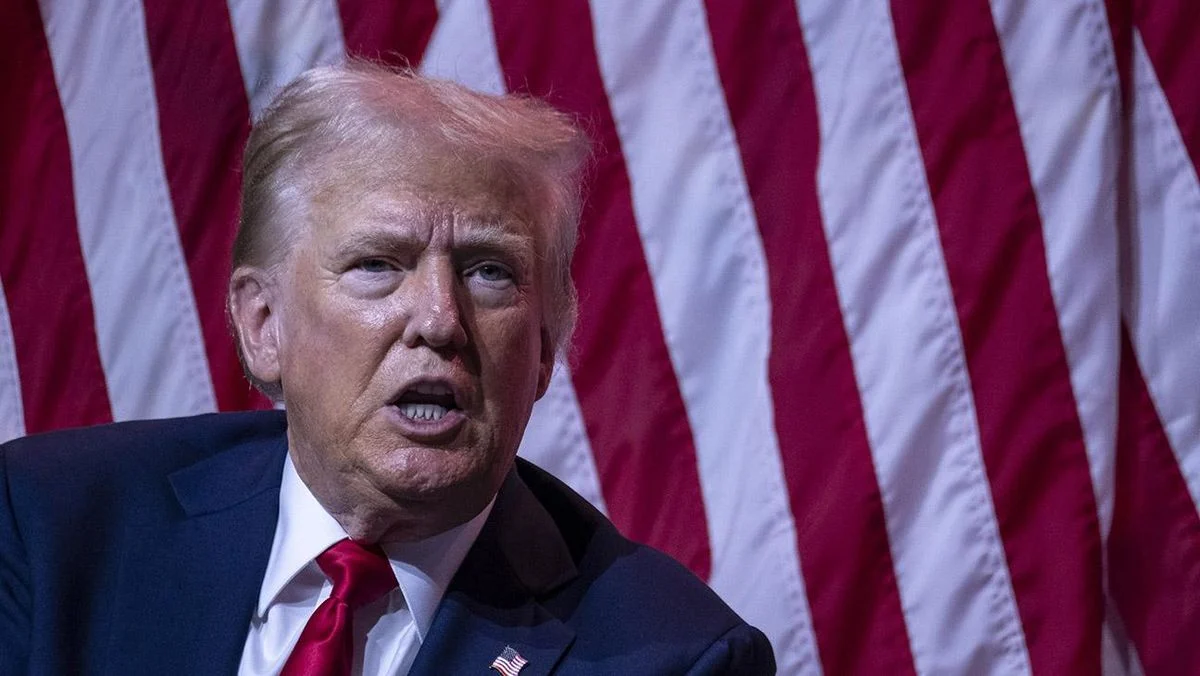 Donald Trump speaking at a conference with the American flag in the background.